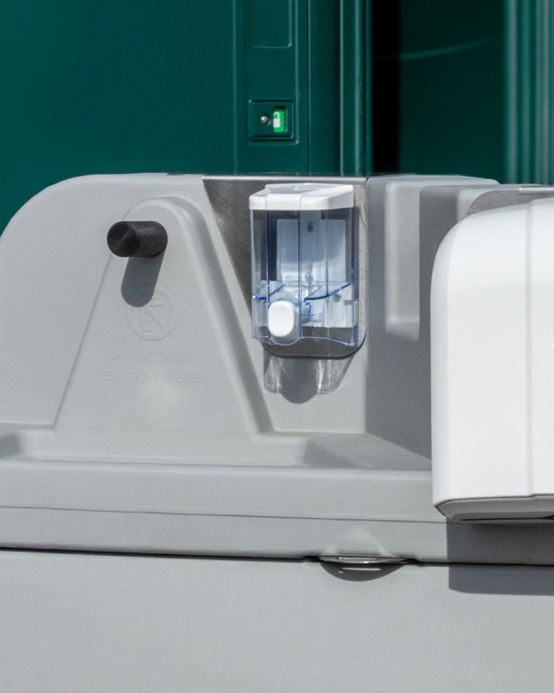 Detailansicht der Handwaschstation: Wasserhahn und Seifenspender in Nahaufnahme, grüne WC-Kabinentür im Hintergrund.