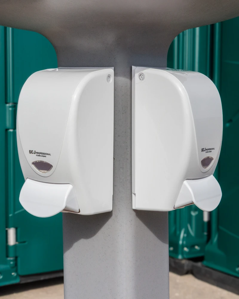 Zwei Desinfektionsmittelspender am s'Örtle-Ständer – Nahaufnahme der Pumpspender mit grünen Toilettenkabinen im Hintergrund.